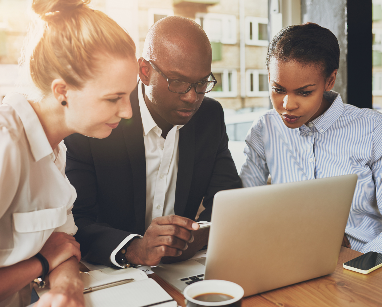 Three colleagues closely collaborating and looking at a laptop screen at a wooden table with a coffee cup and notebook.