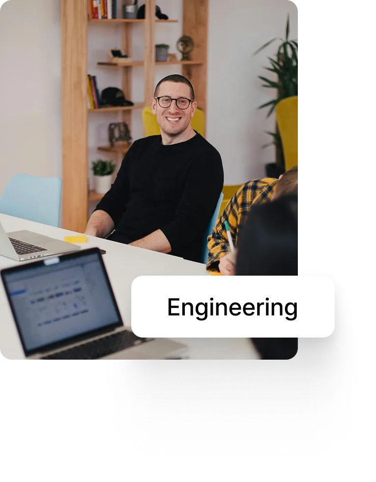 Smiling man with glasses sitting at a table with laptops in a modern office.