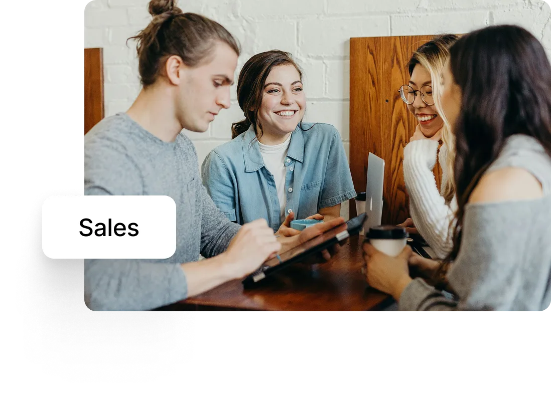 Four young people sitting around a table, smiling and engaged in a discussion with digital devices and coffee cups.
