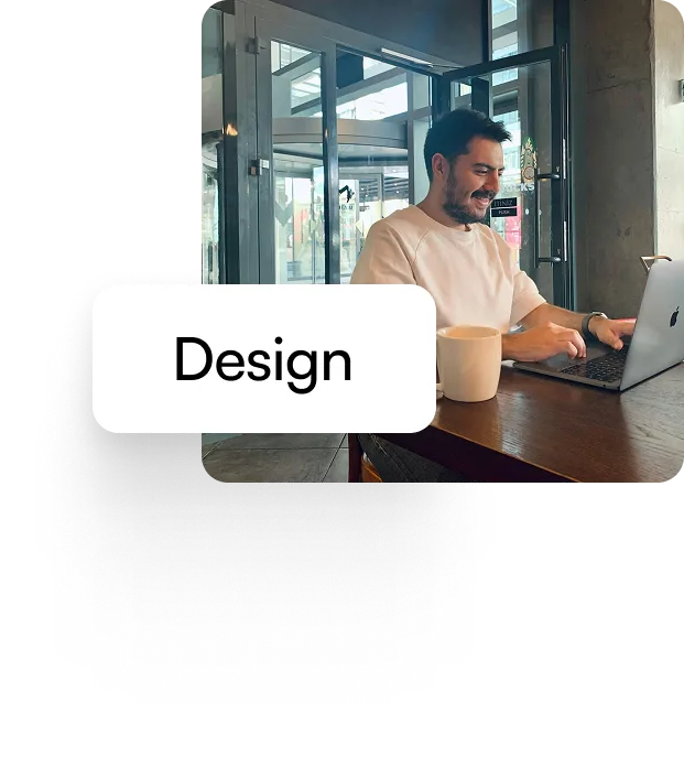 Man smiling and working on a laptop at a wooden table in a modern cafe with a coffee cup nearby.