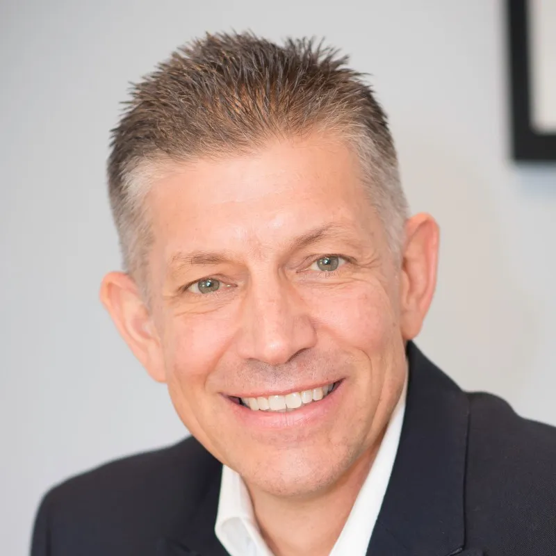 Smiling middle-aged man with short gray hair wearing a dark blazer and white shirt.