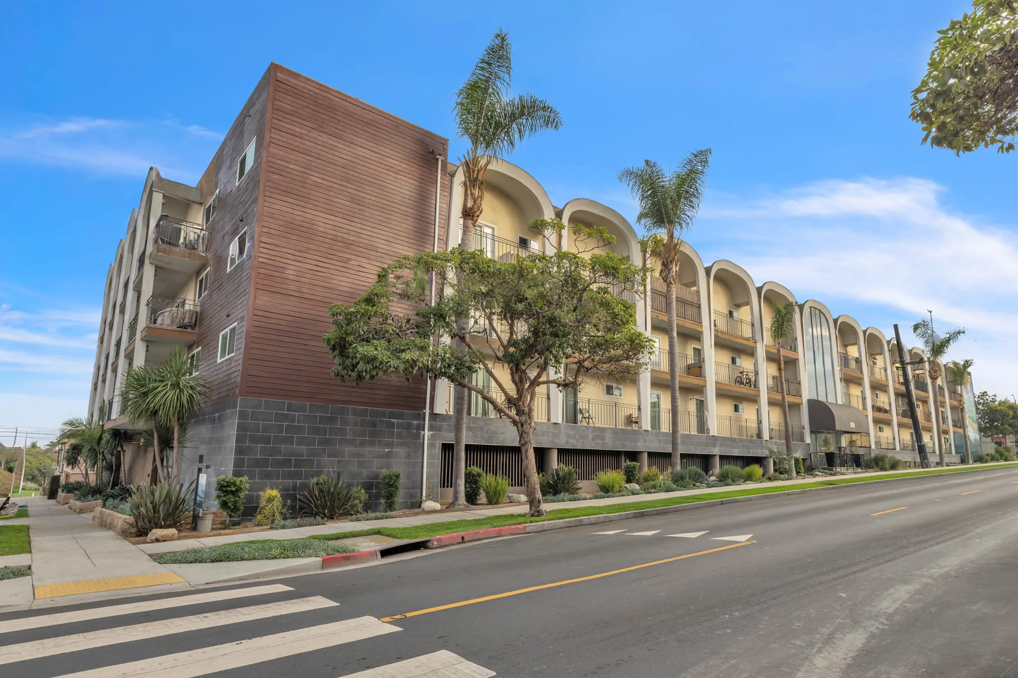 Modern four-story apartment building with balconies, palm trees, and a crosswalk in front under a clear blue sky.