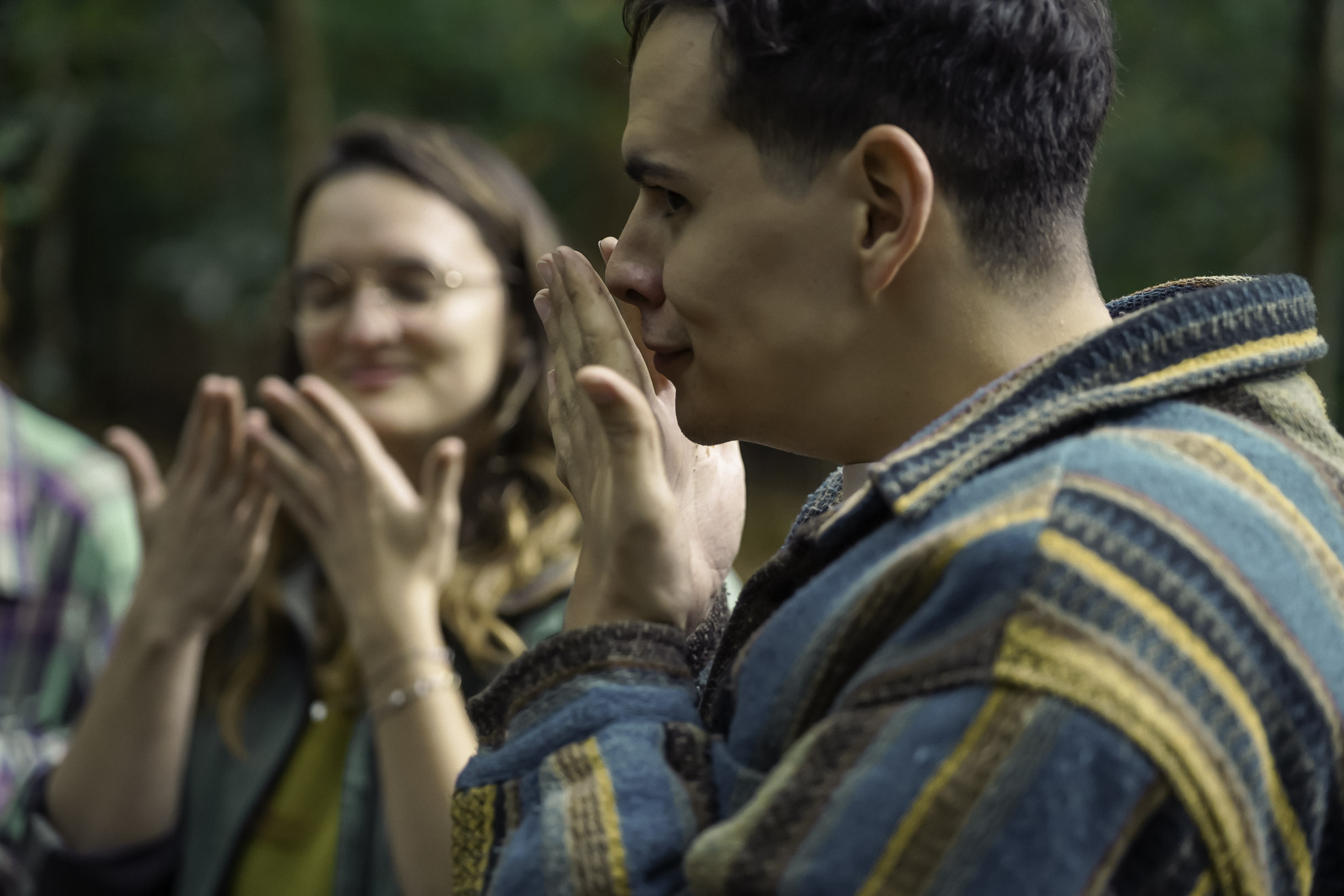 Close-up of a man wearing a striped jacket holding his hands near his face, with a smiling woman clapping blurred in the background.