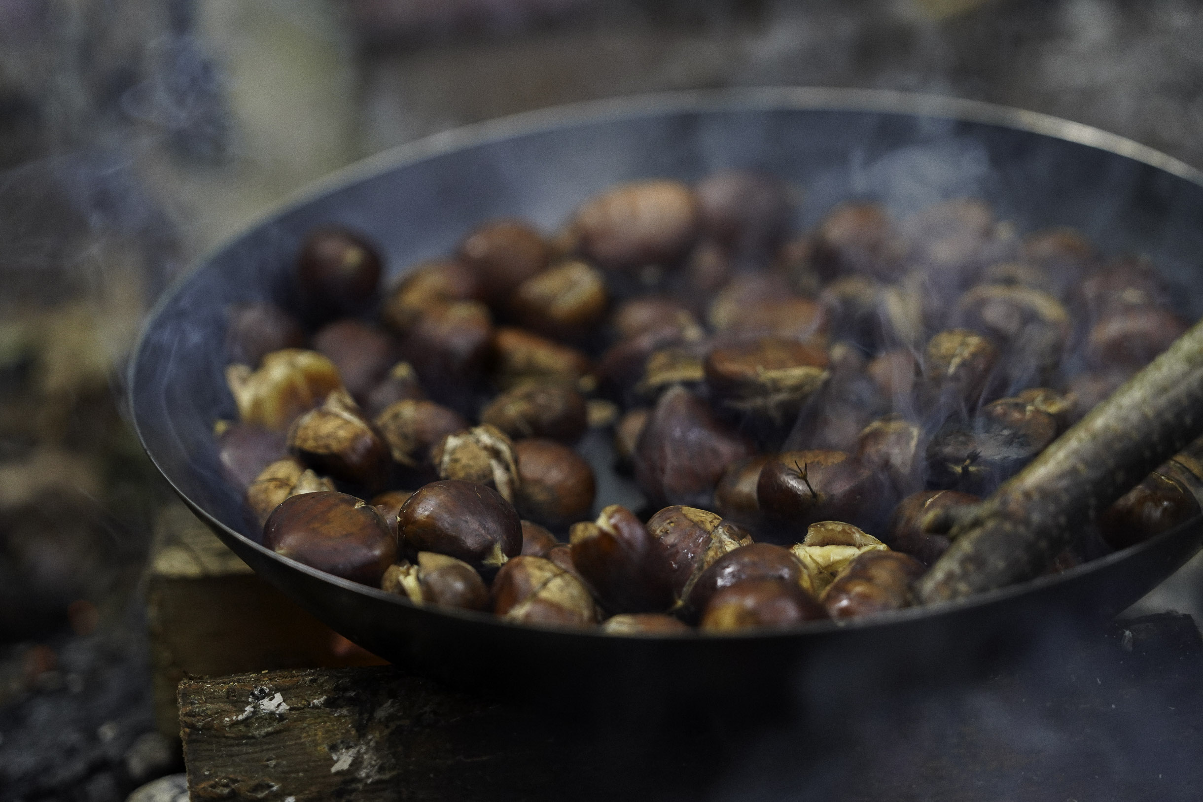 Chestnuts roasting in a pan over an open wood fire with smoke rising.