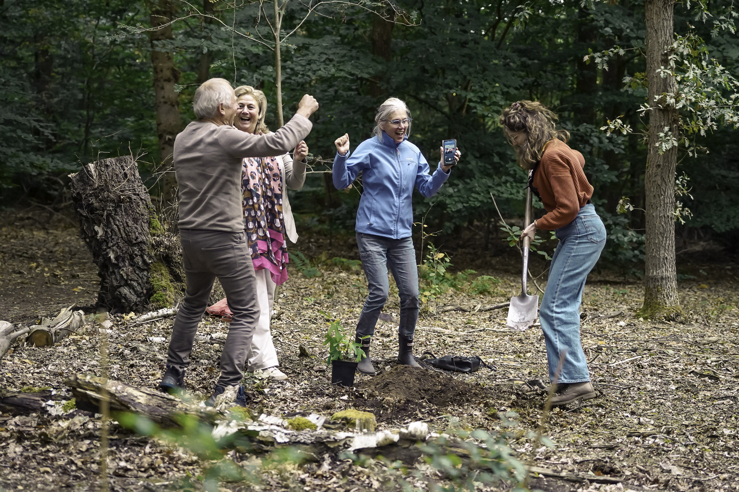Four adults outdoors in a forest, one woman holding a shovel digging soil, another holding a smartphone, and two others celebrating beside a small tree ready for planting.