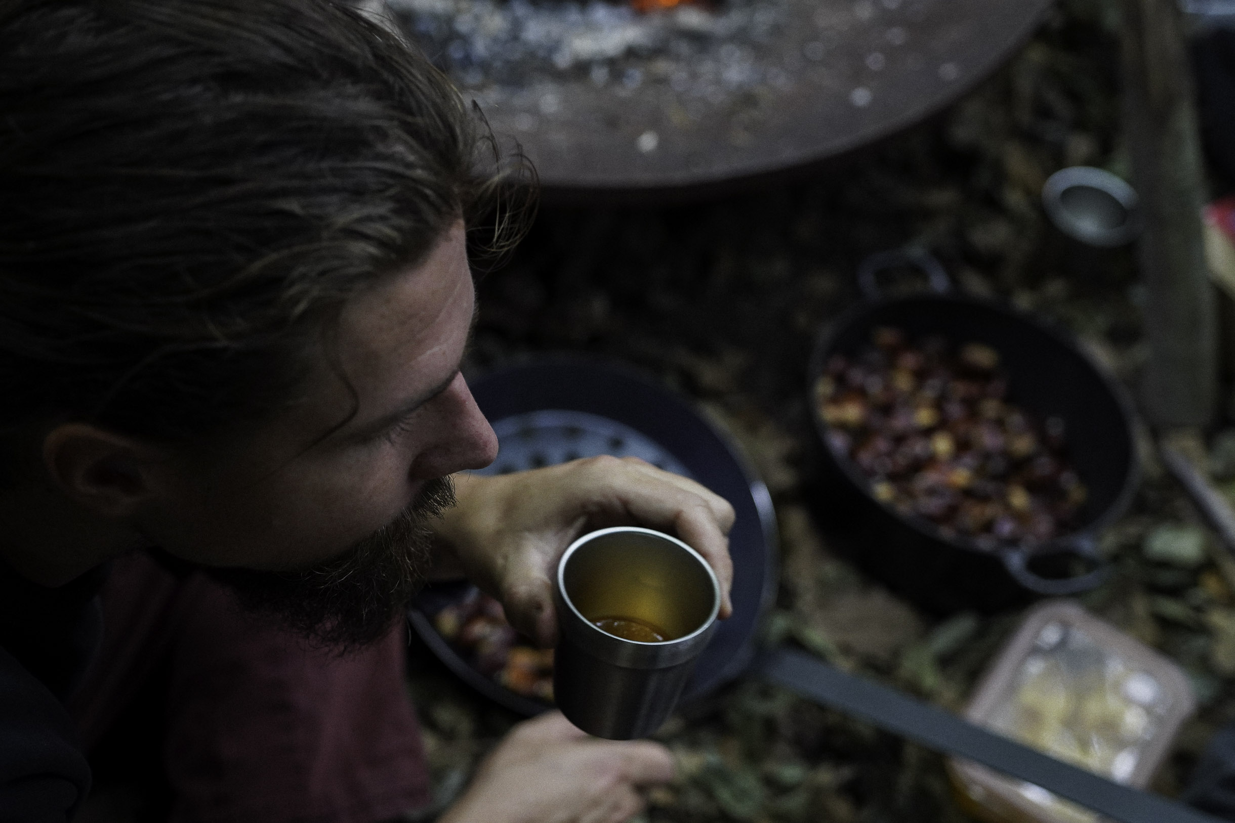 Man with beard holding a metal cup with liquid, with containers of coffee cherries in the background outdoors.