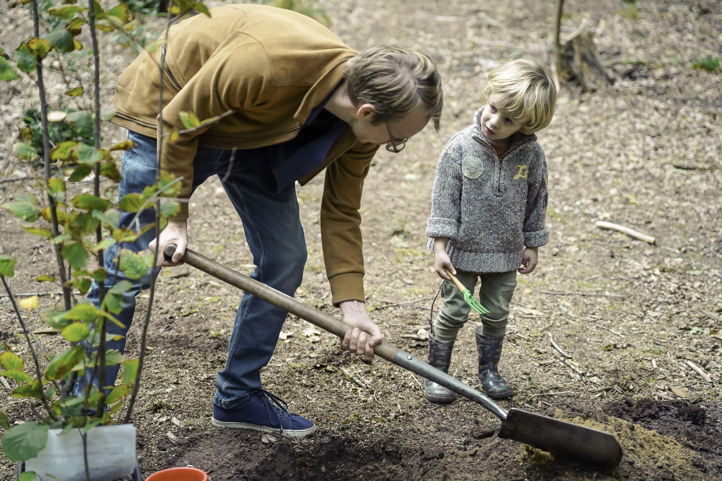 Man digging a hole in the ground while a young child watches holding a small green gardening tool in a wooded area.
