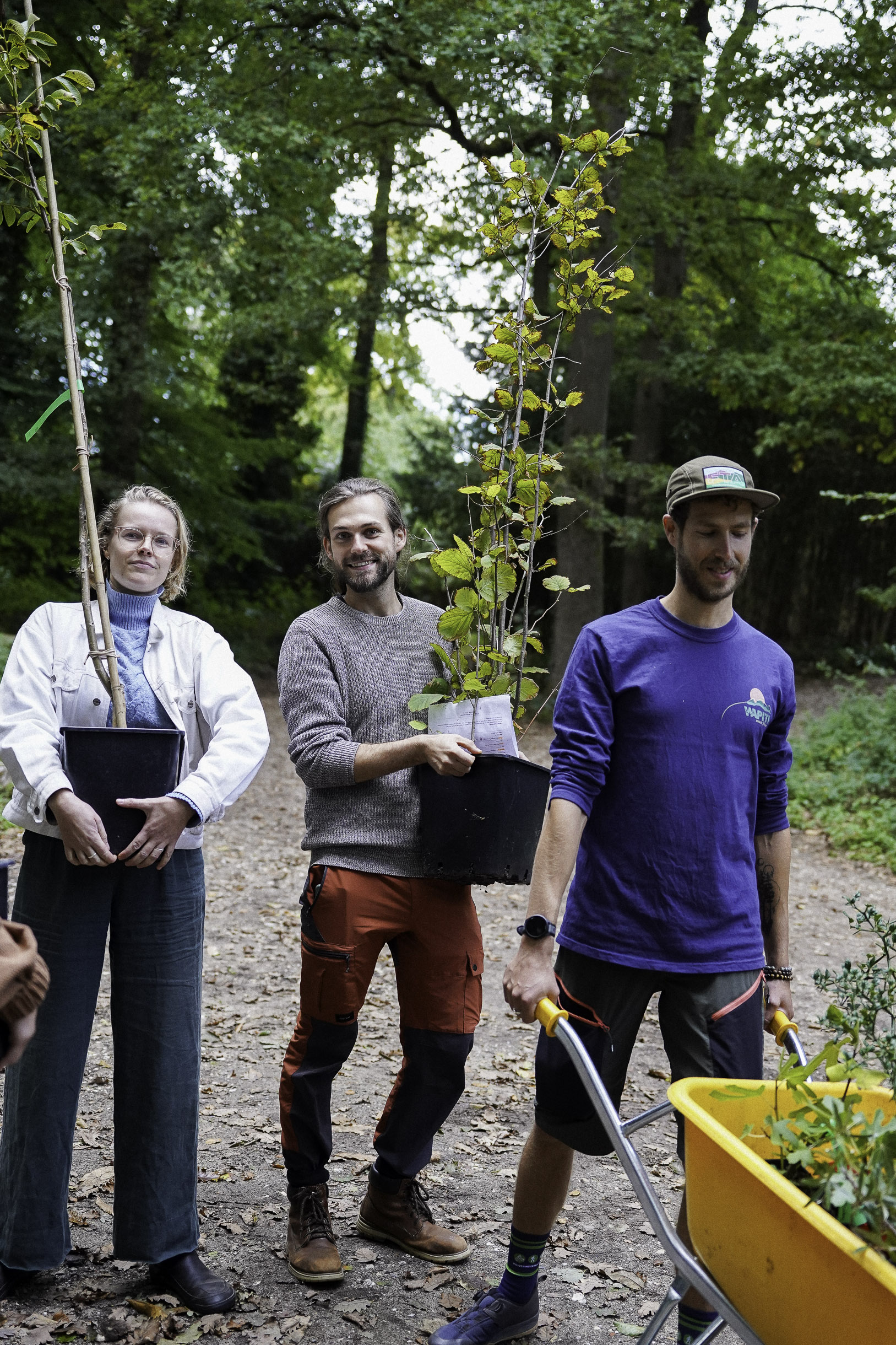 Three people outdoors holding young potted trees and a yellow wheelbarrow on a forest path.