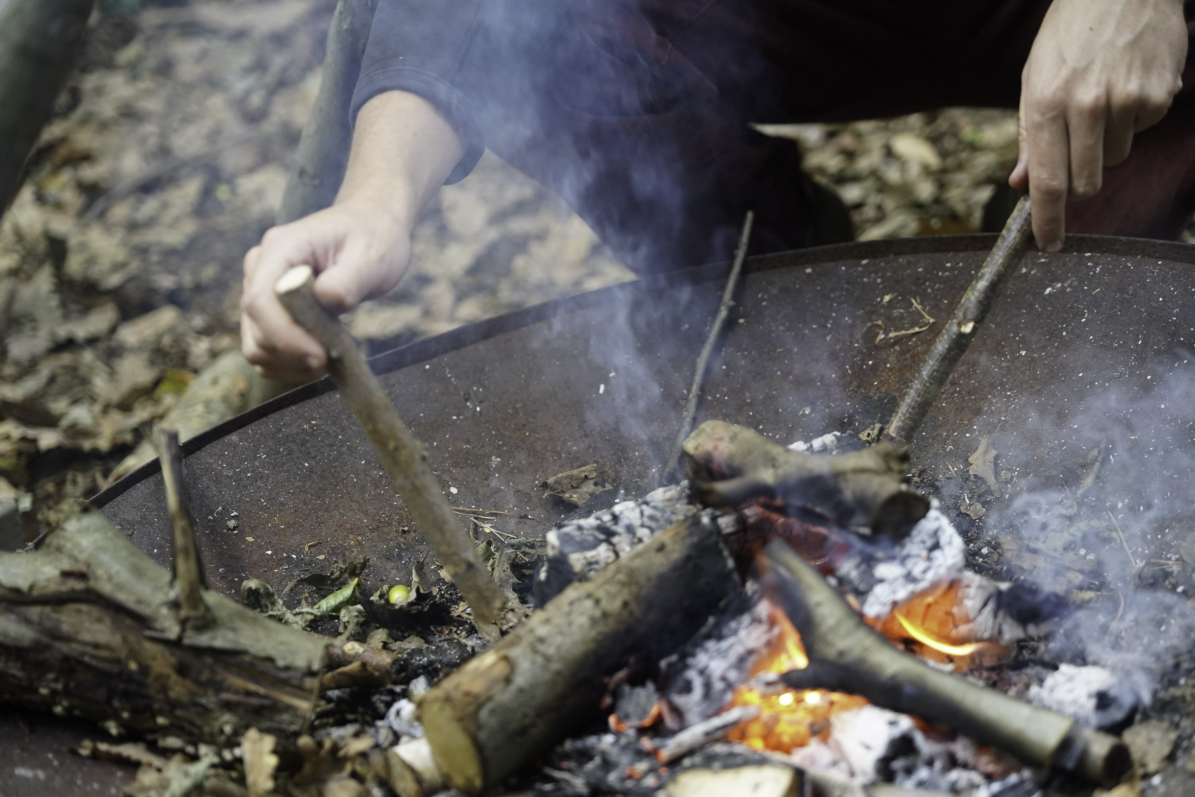 Hands using sticks to tend a smoky campfire with burning logs and embers in an outdoor setting.