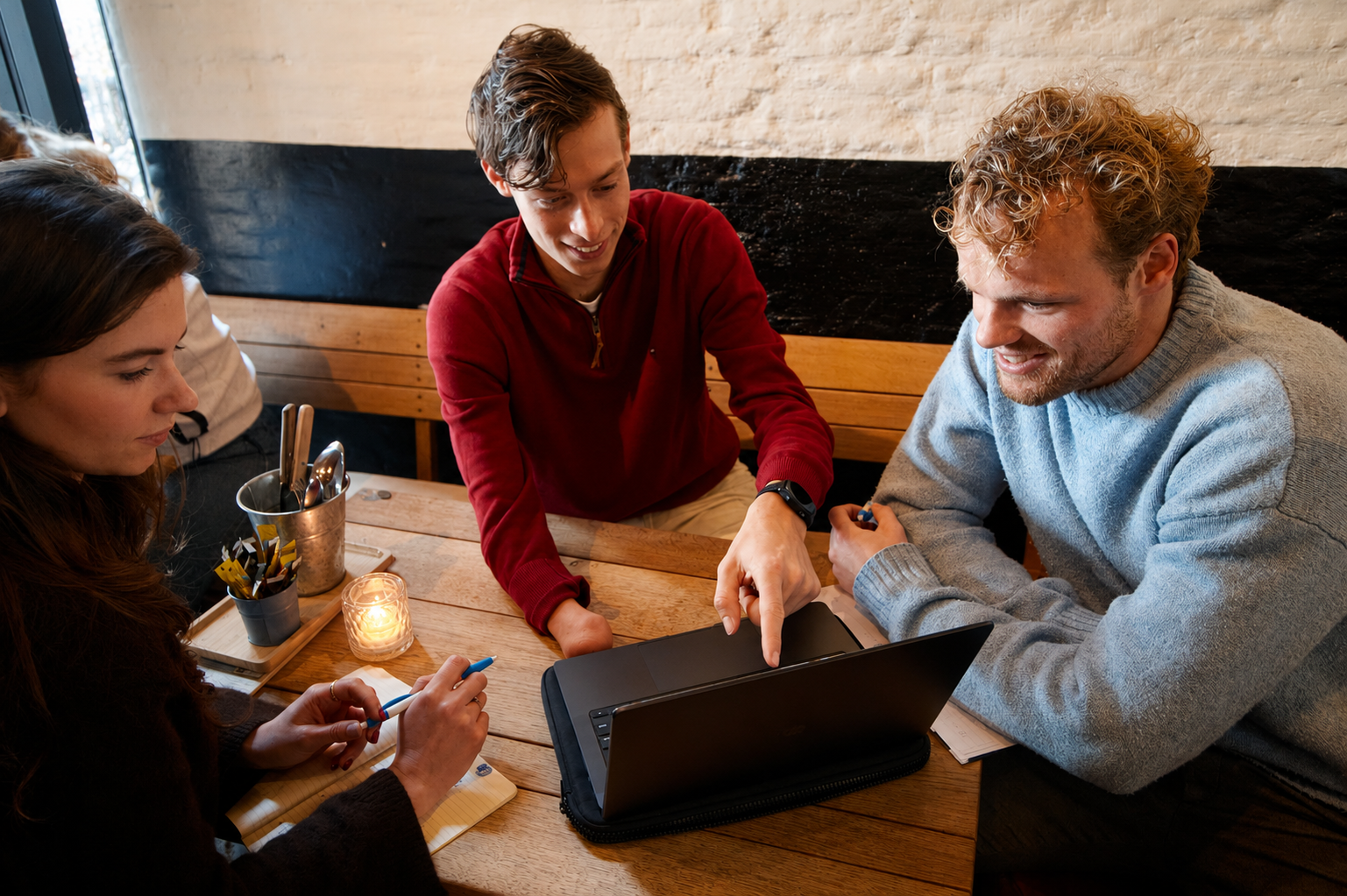 Three young adults brainstorming at a wooden table, looking at a laptop screen while one person points at it and another takes notes.