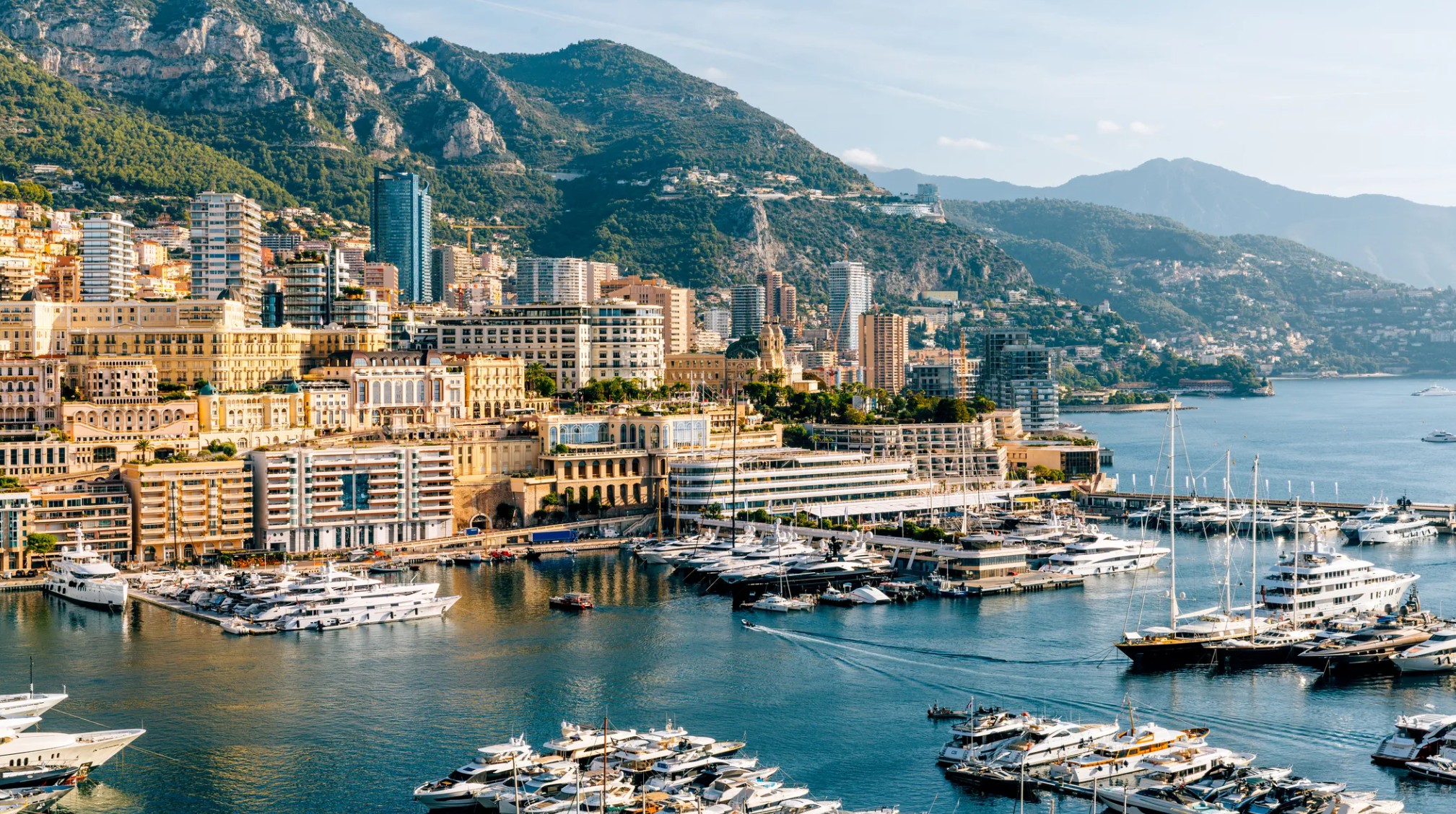 View of Monaco harbor filled with yachts, surrounded by modern buildings and green mountains under a clear sky.
