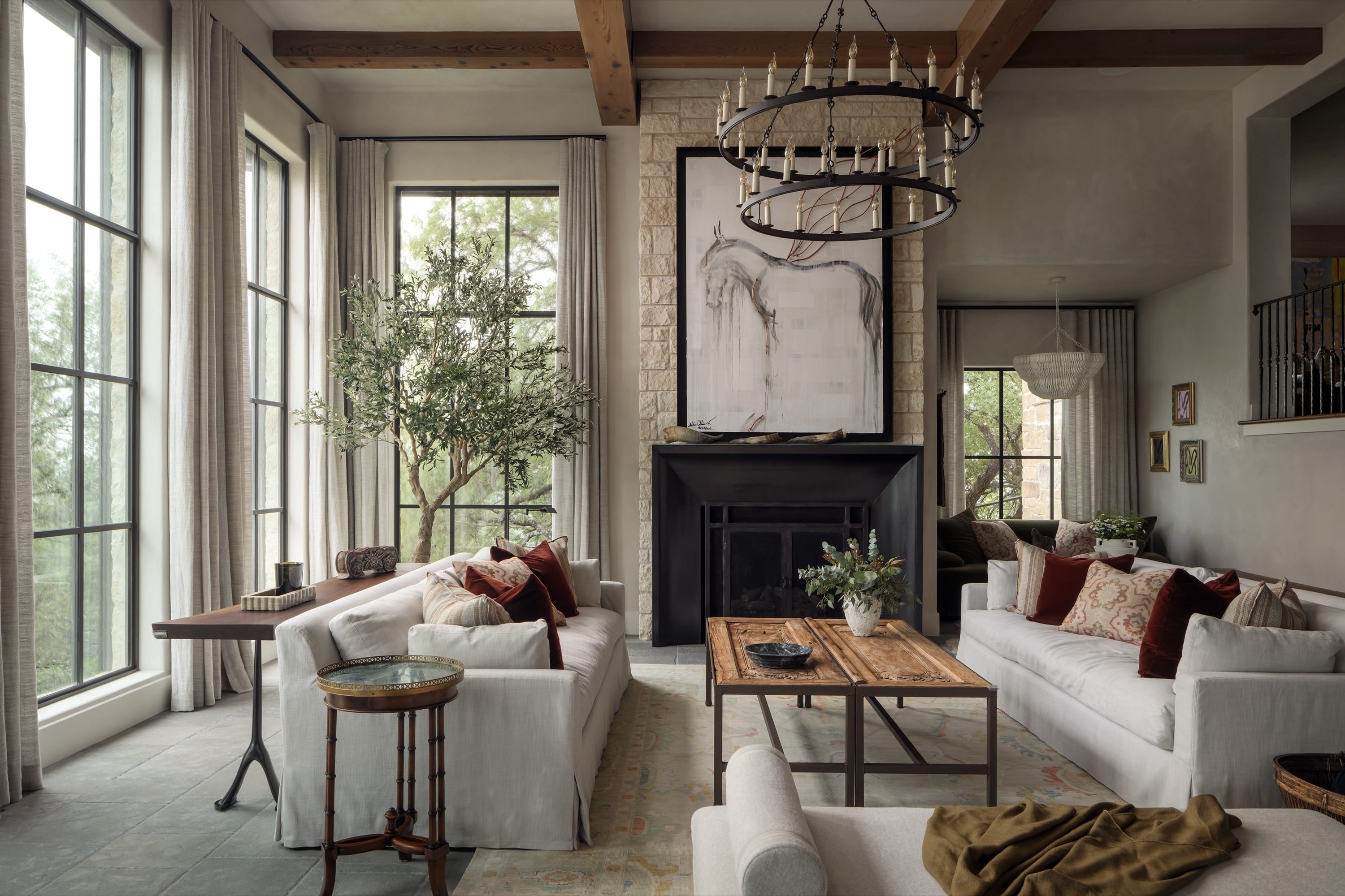 Modern living room with two white sofas adorned with red and patterned pillows, a wooden coffee table, a fireplace with horse artwork above, large windows with beige curtains, and a green leafy plant.