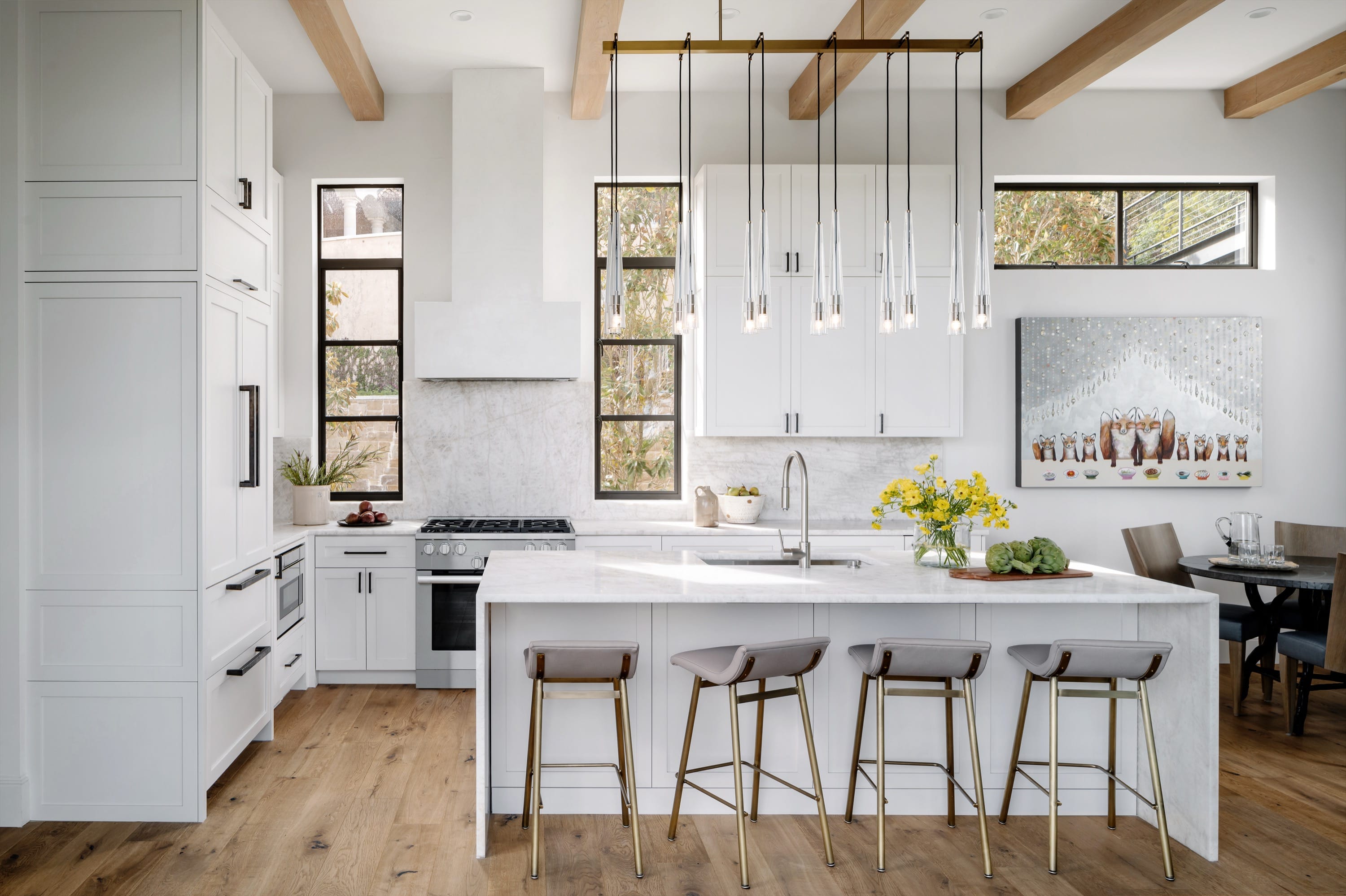 Bright modern kitchen with white cabinetry, marble island with four stools, wooden floor, pendant lighting, and artwork on wall.