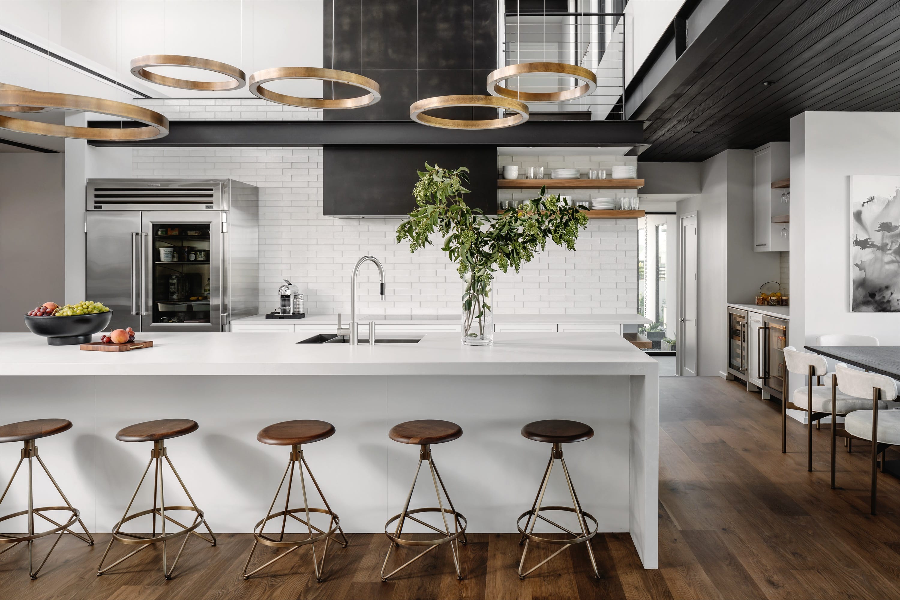 Modern kitchen with white island, five wooden stools, circular pendant lights, stainless steel refrigerator, and a vase with green foliage on the island.
