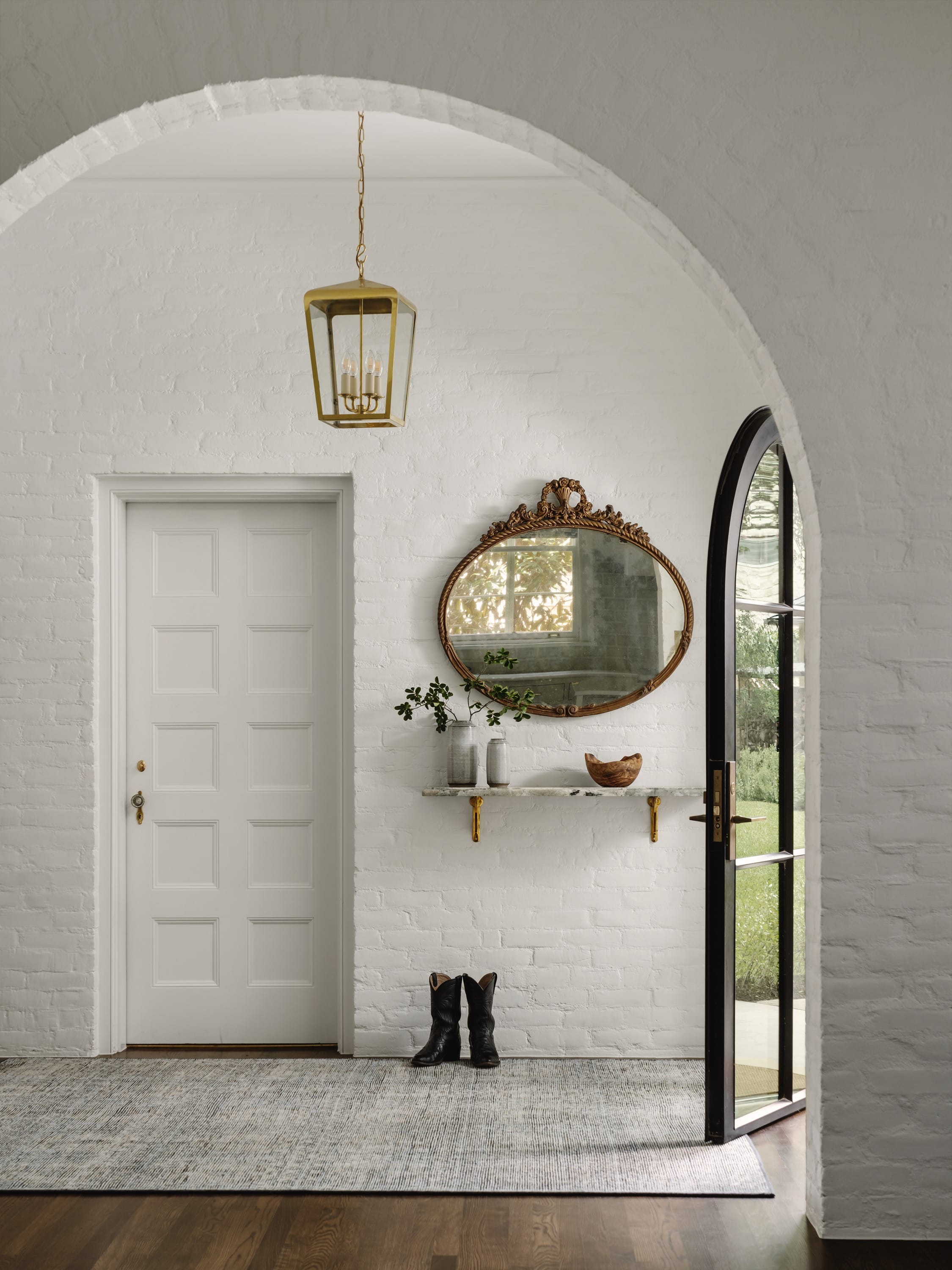 Entryway with white brick walls, an arched doorway with a black-framed glass door open to a garden, white paneled door, ornate oval mirror above a marble shelf holding vases and a wooden bowl, and pair of black boots on a gray rug.