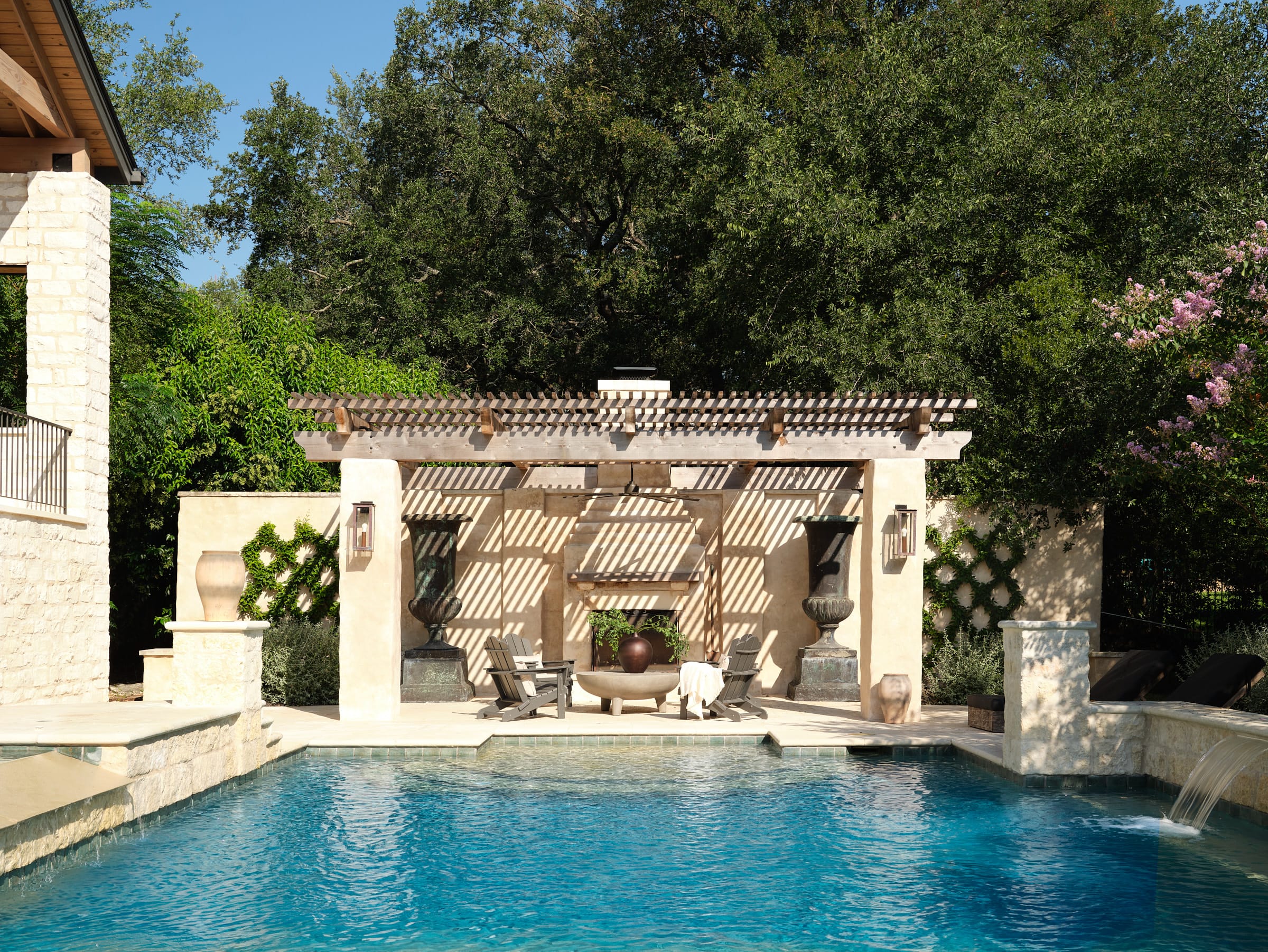 Outdoor pool with stone deck, pergola-covered seating area with chairs and a fire pit, surrounded by lush green trees.