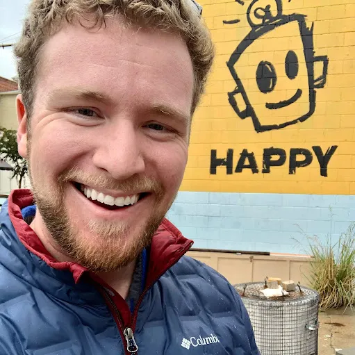 A man smiling in front of a yellow wall with a happy sign.