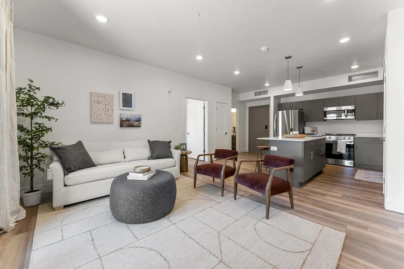 Modern open living room with light gray sofa, two burgundy armchairs, decorative ottoman, and kitchen with gray cabinetry and stainless steel appliances.