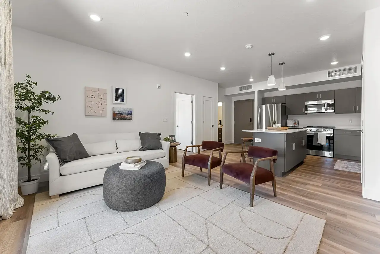 Modern open living room with light gray sofa, two burgundy armchairs, decorative ottoman, and kitchen with gray cabinetry and stainless steel appliances.