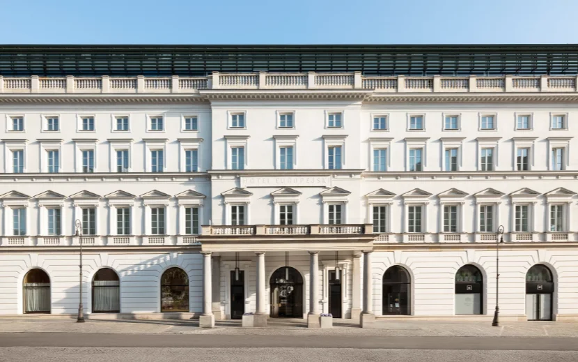 Symmetrical white neoclassical building facade with multiple windows, central portico entrance, and two street lamps on a clear day.