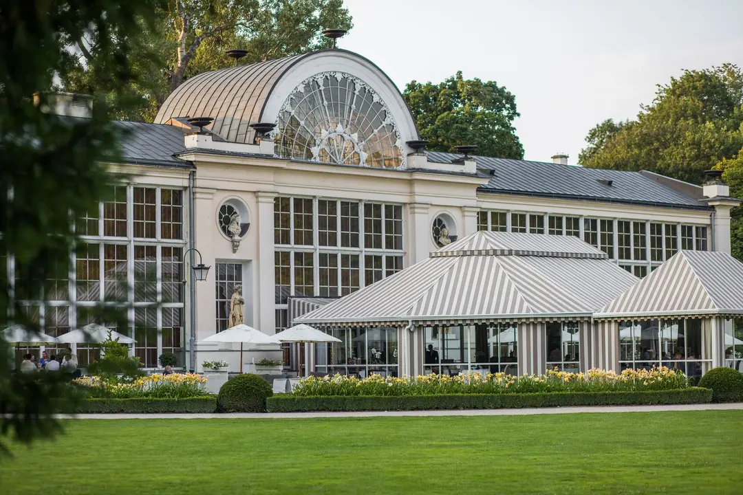 Elegant historical building with large arched windows and a striped tented outdoor seating area, surrounded by greenery and manicured lawns.