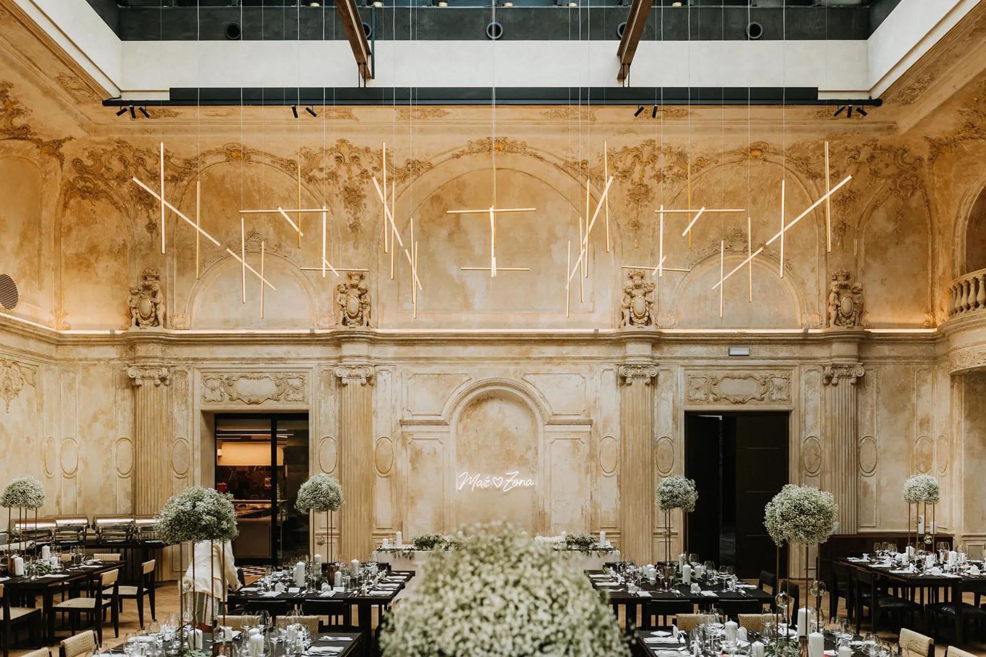 Elegant wedding reception setup in a historic venue with ornate walls, modern pendant lights, tall floral centrepieces, and neatly arranged tables.