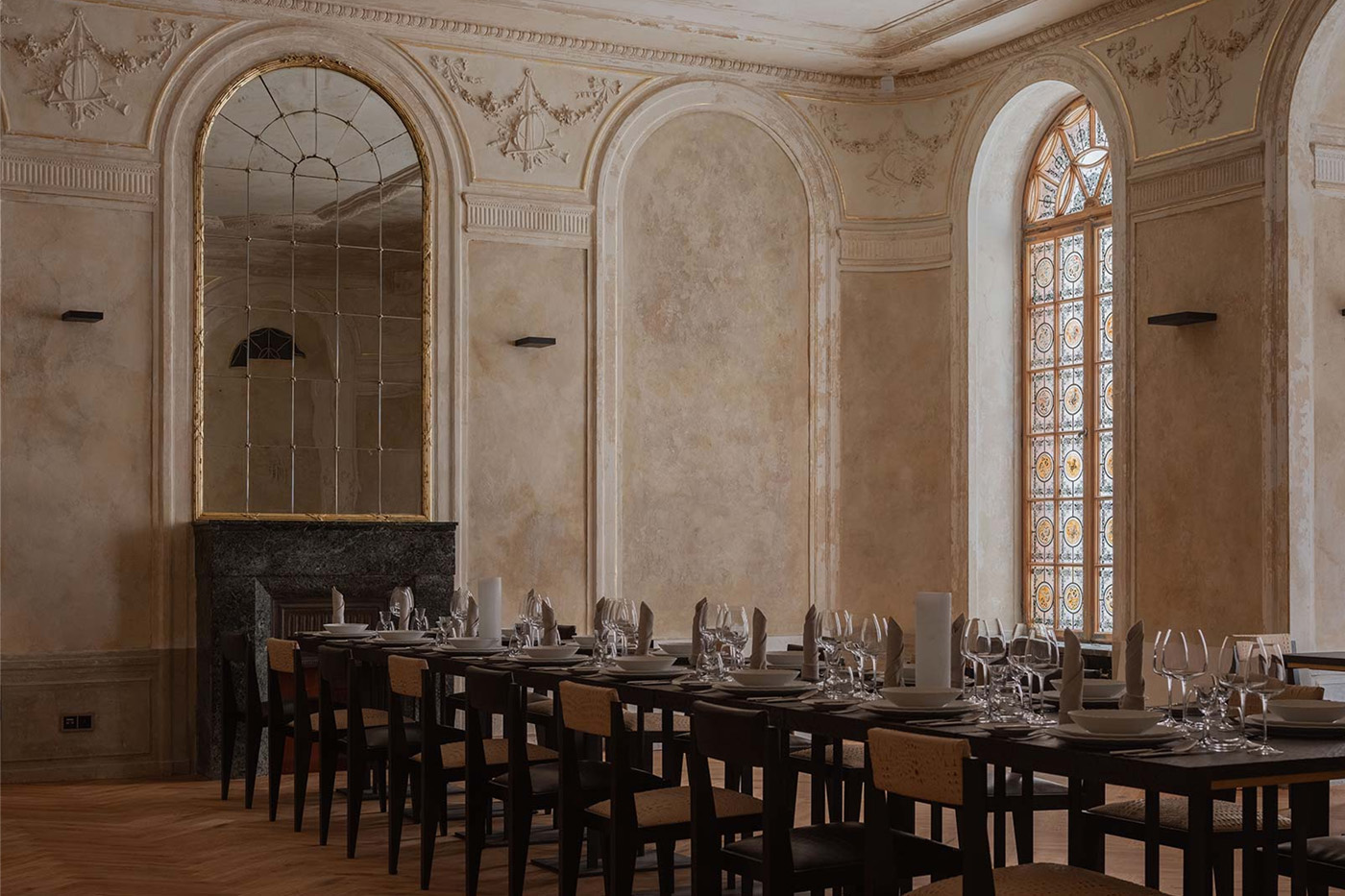 Elegant dining room with long tables set with plates, glasses, and napkins, featuring antique arched mirror and stained glass window.
