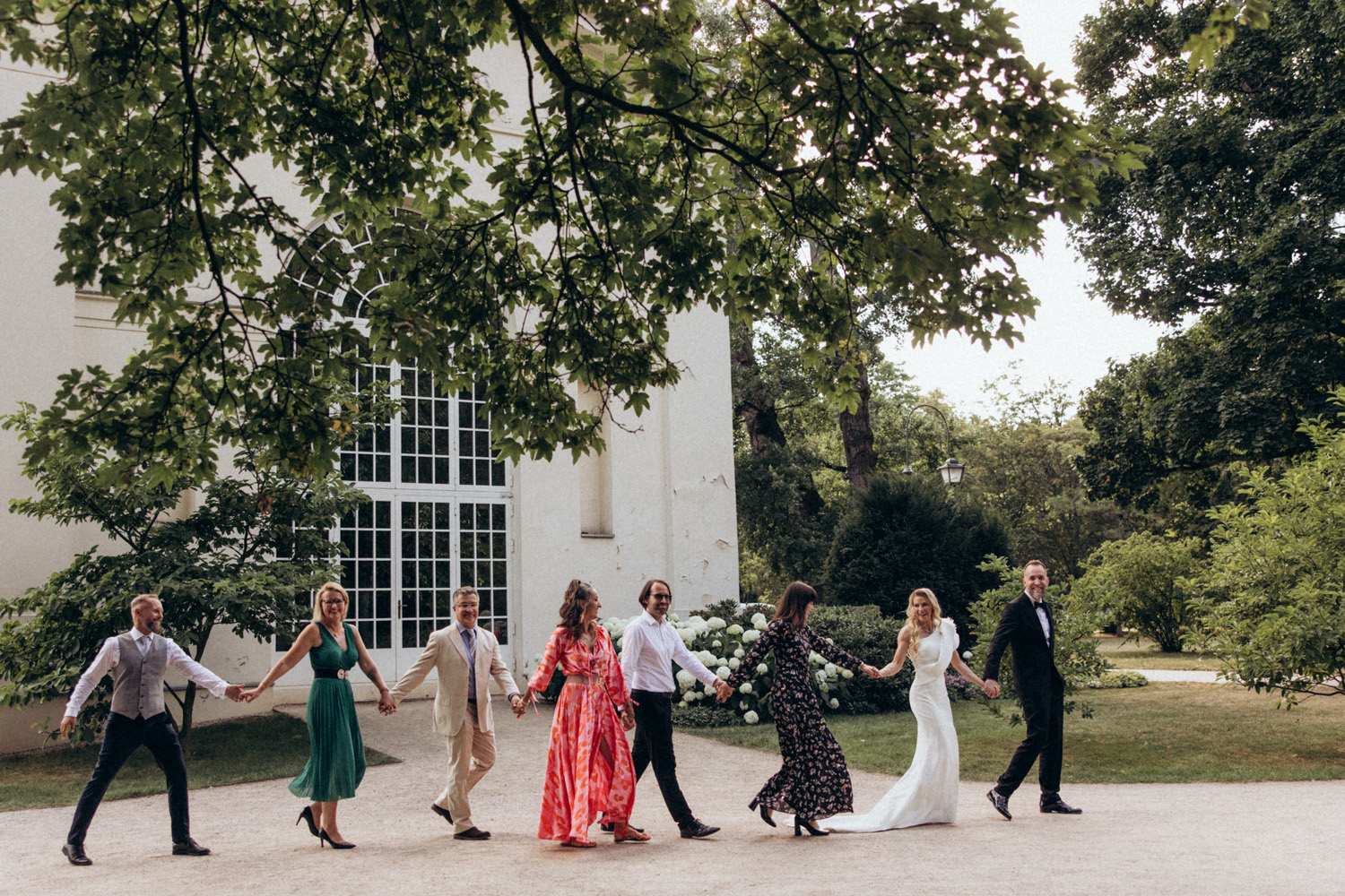 Seven people holding hands walking outdoors near a large windowed building surrounded by trees and bushes.