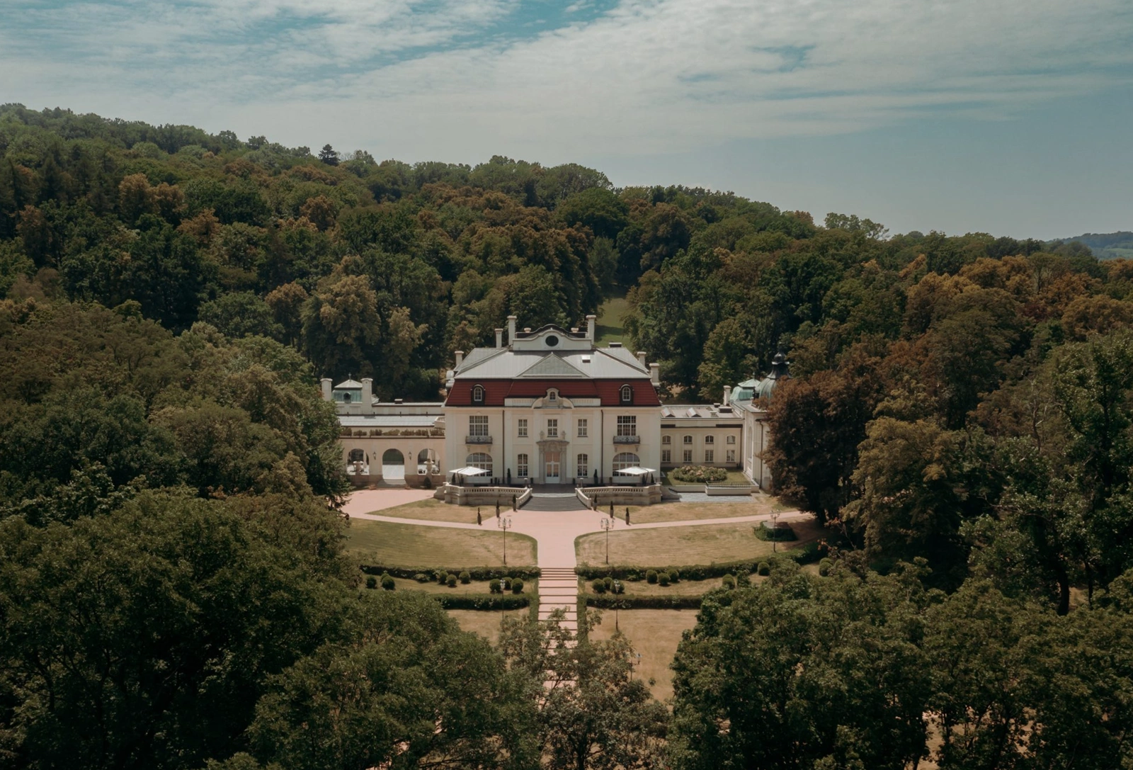 Large classical mansion with a red roof surrounded by dense green forest under a partly cloudy sky.