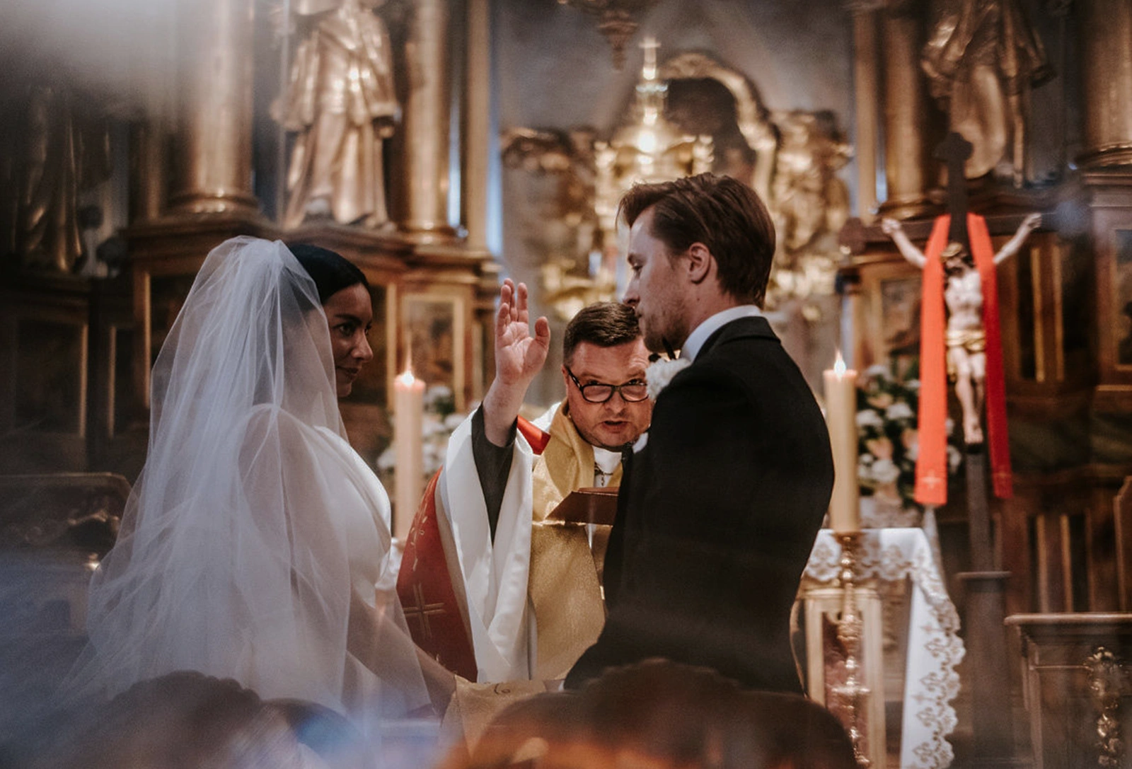 Woman in a glittery white gown with long wavy hair standing at a bathroom sink, looking into an ornate oval mirror.