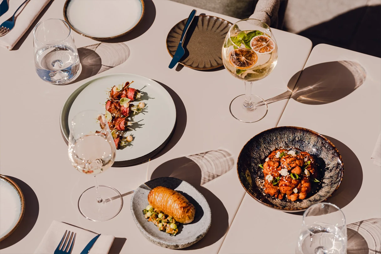 Table set with gourmet dishes including a plate of rolled meat with garnish, a Hasselback potato, a bowl of cauliflower with sauce, and glasses of white wine and water.