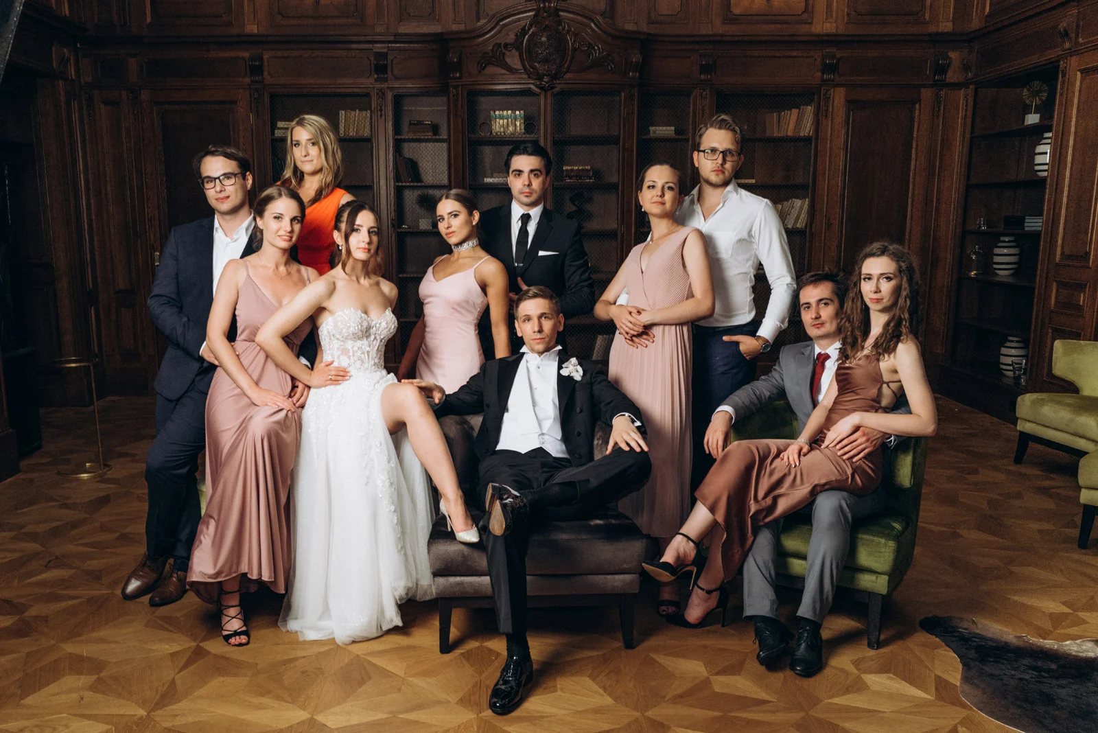 Group portrait of elegantly dressed men and women posing in a wood-paneled room, featuring a bride in a white gown and a groom in a tuxedo seated at the centre.