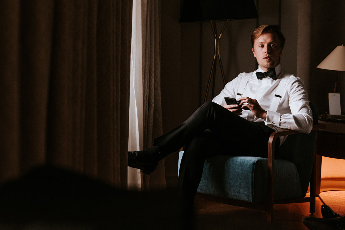 Young man in formal white shirt, black bow tie, and suspenders sitting in a blue armchair holding a phone.
