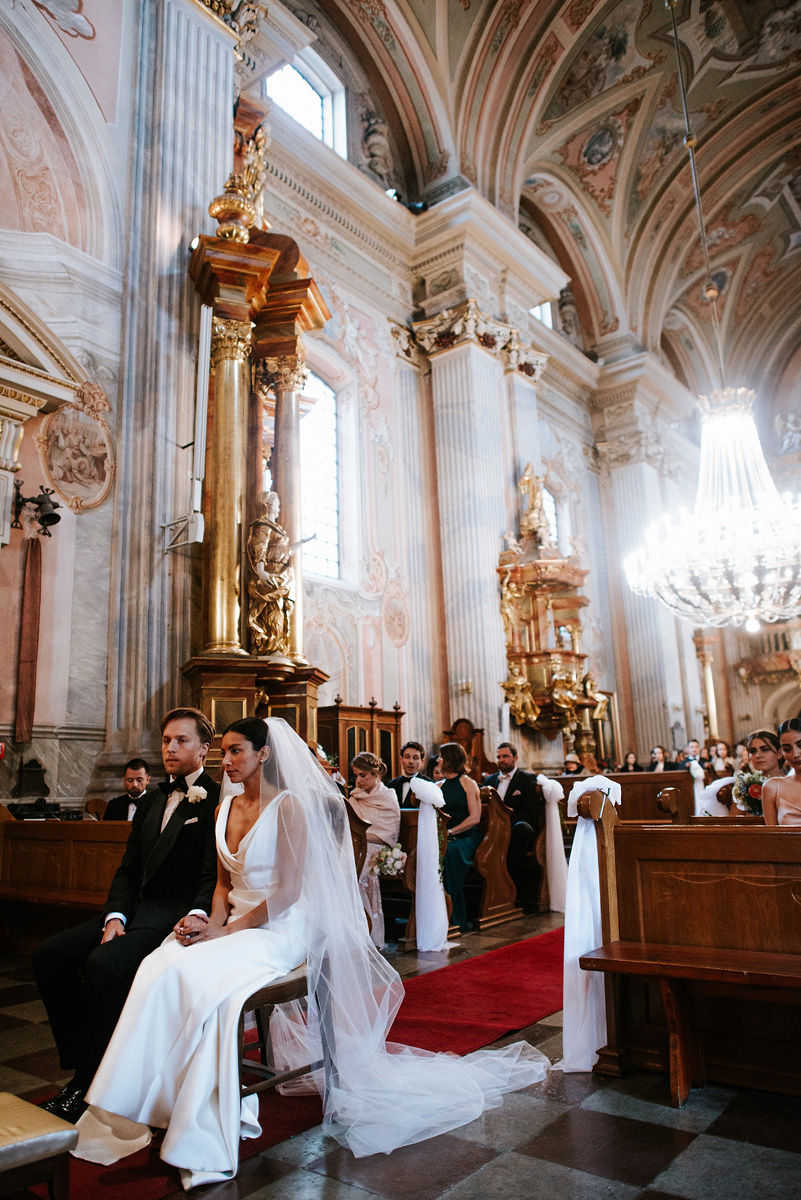 Bride in white dress and veil and groom in black tuxedo sitting together in ornately decorated church during wedding ceremony.