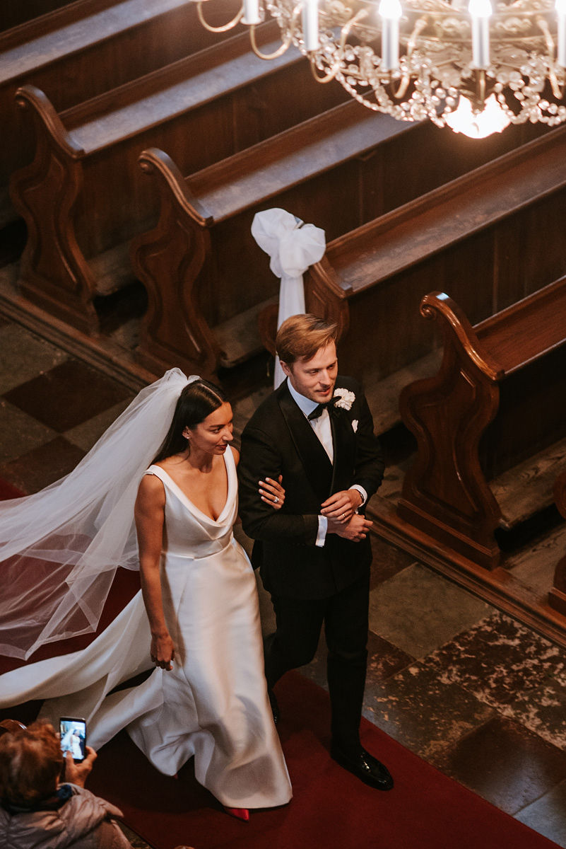 Bride in a white gown and veil walking arm-in-arm with groom in a black tuxedo down a church aisle with wooden pews and a chandelier above.