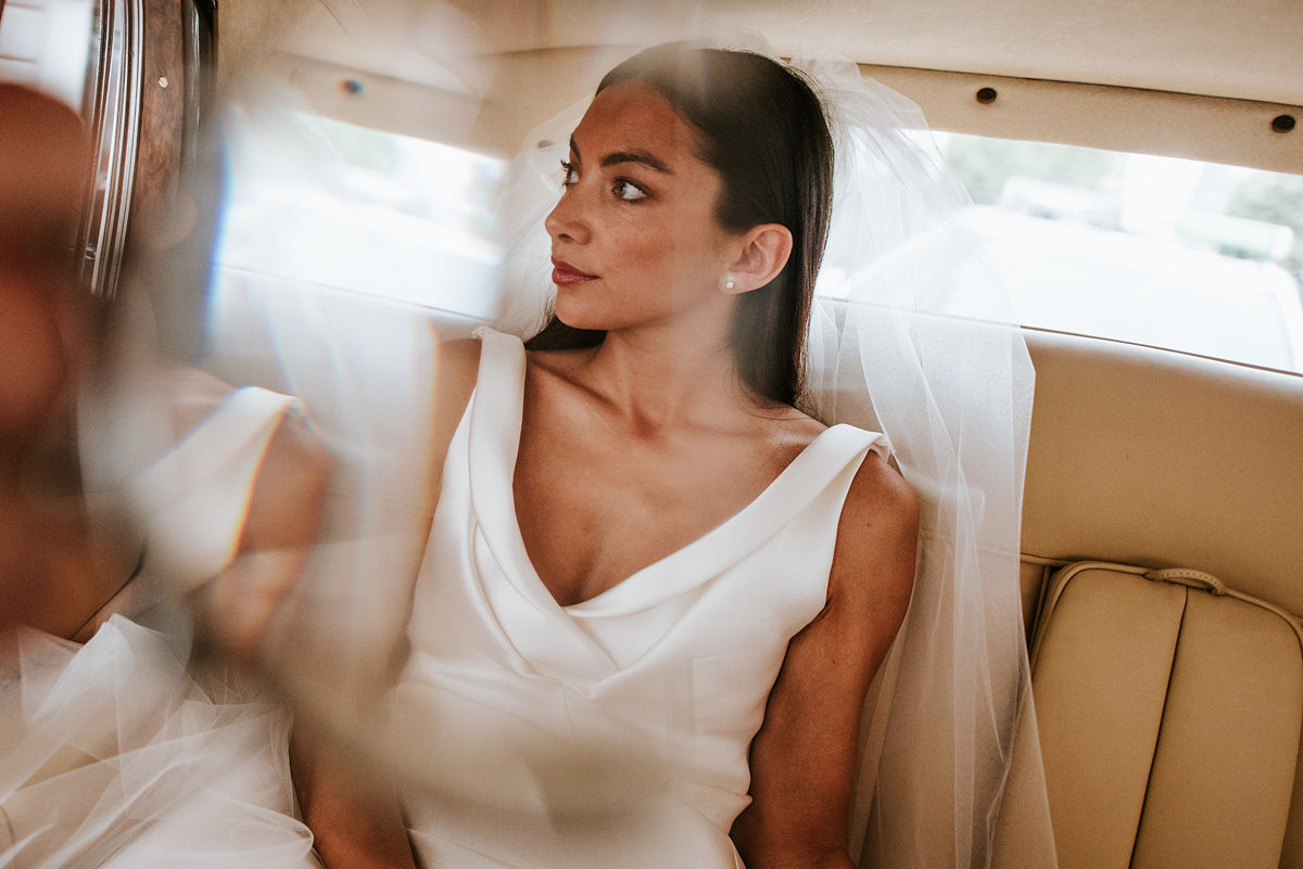 Bride in a white wedding dress and veil sitting in the back seat of a car, looking to the side.