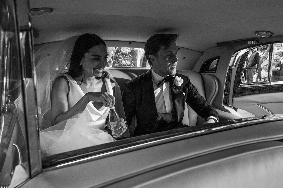 Black and white photo of a smiling bride in a wedding dress and veil holding a bottle, sitting next to a groom in a tuxedo inside a vintage car.