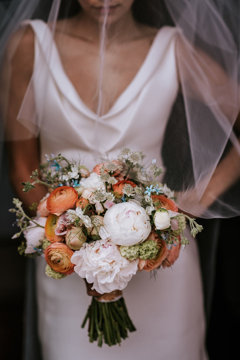 Bride in white dress holding a bouquet of white, peach, and green flowers under a veil.