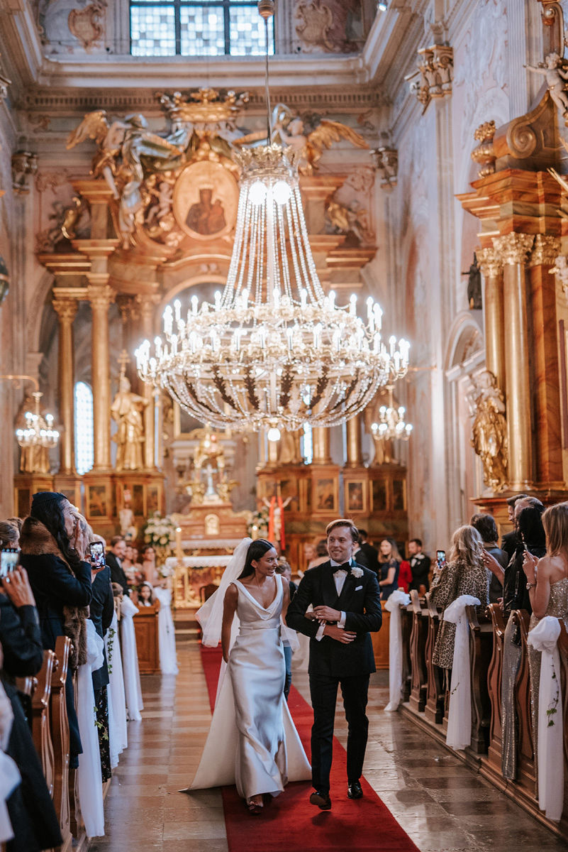Bride and groom walking down the aisle of an ornately decorated church with a large chandelier overhead.