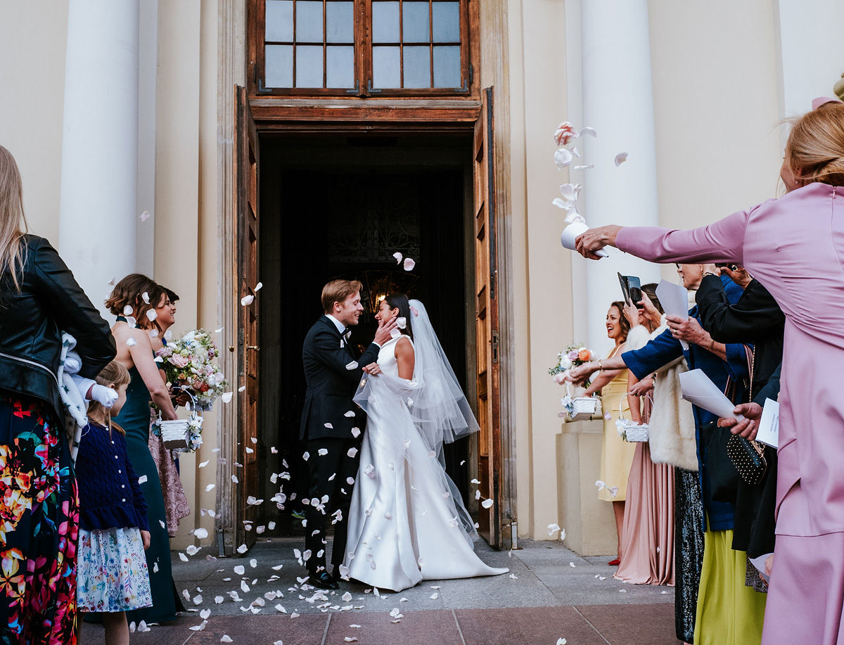 Bride and groom kiss outside church door while guests throw white flower petals.