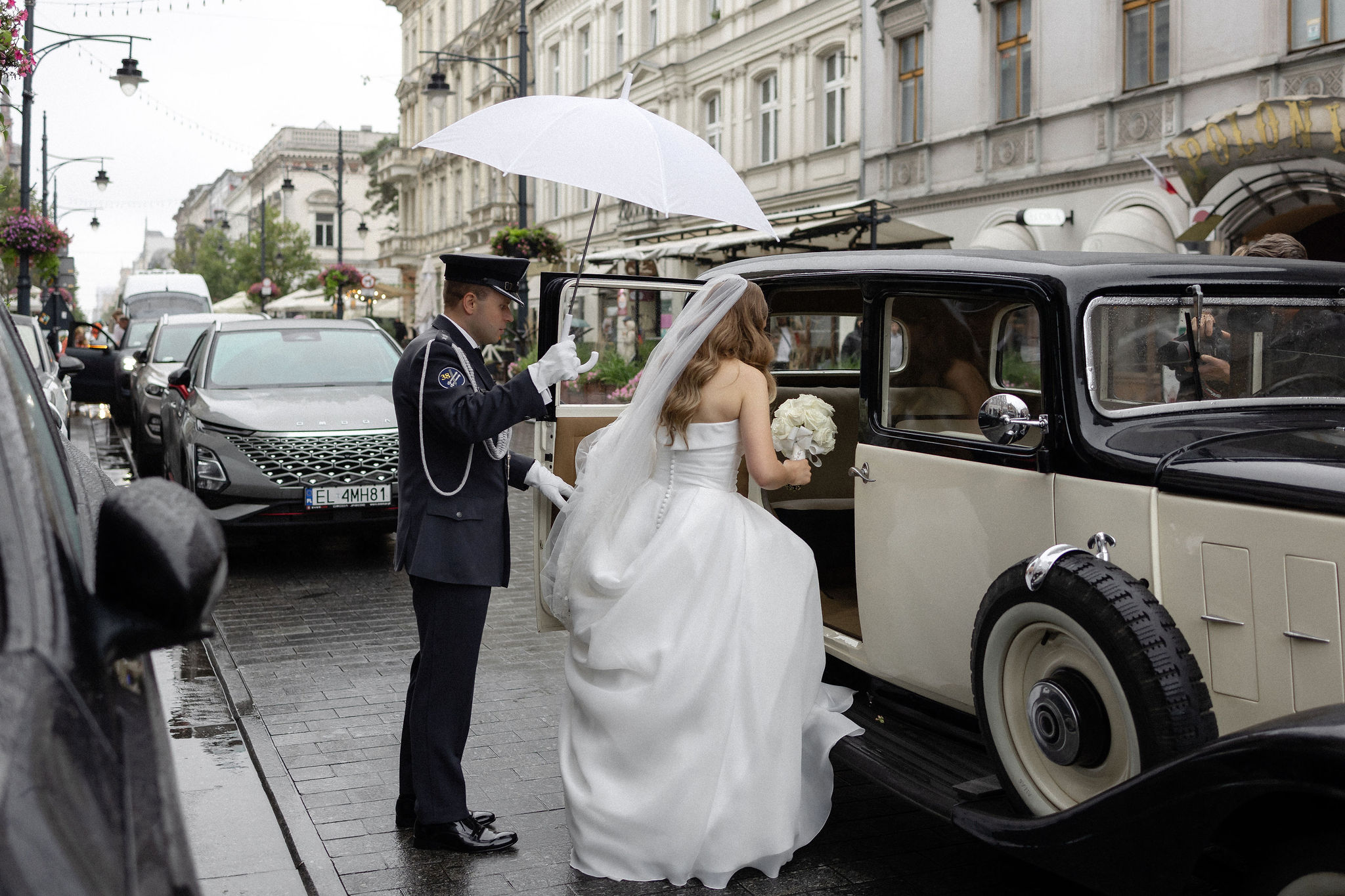 A bride in a white dress with a bouquet entering a vintage car while a chauffeur in uniform holds a white umbrella over her on a rainy city street.