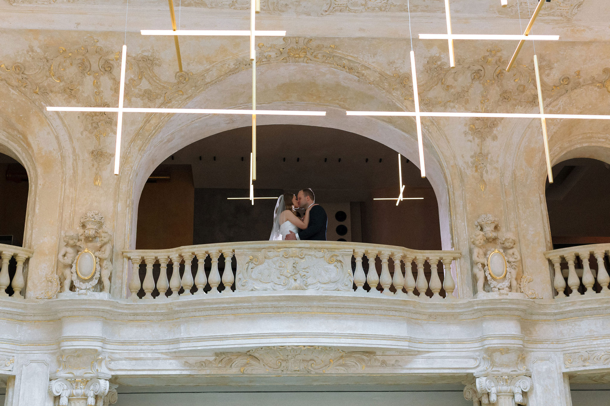 Bride and groom kissing on an ornate balcony under modern cross-shaped ceiling lights.