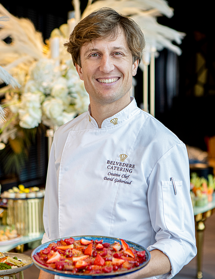 Smiling male chef in white uniform holding a plate of strawberry dessert in a decorated catering setting.