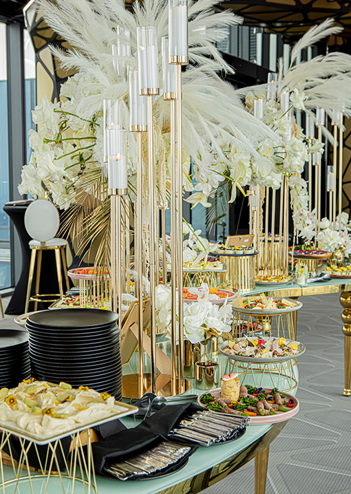 Elegant buffet table with black plates, silverware, and an array of hors d'oeuvres, decorated with tall gold candle holders and white floral arrangements with pampas grass.