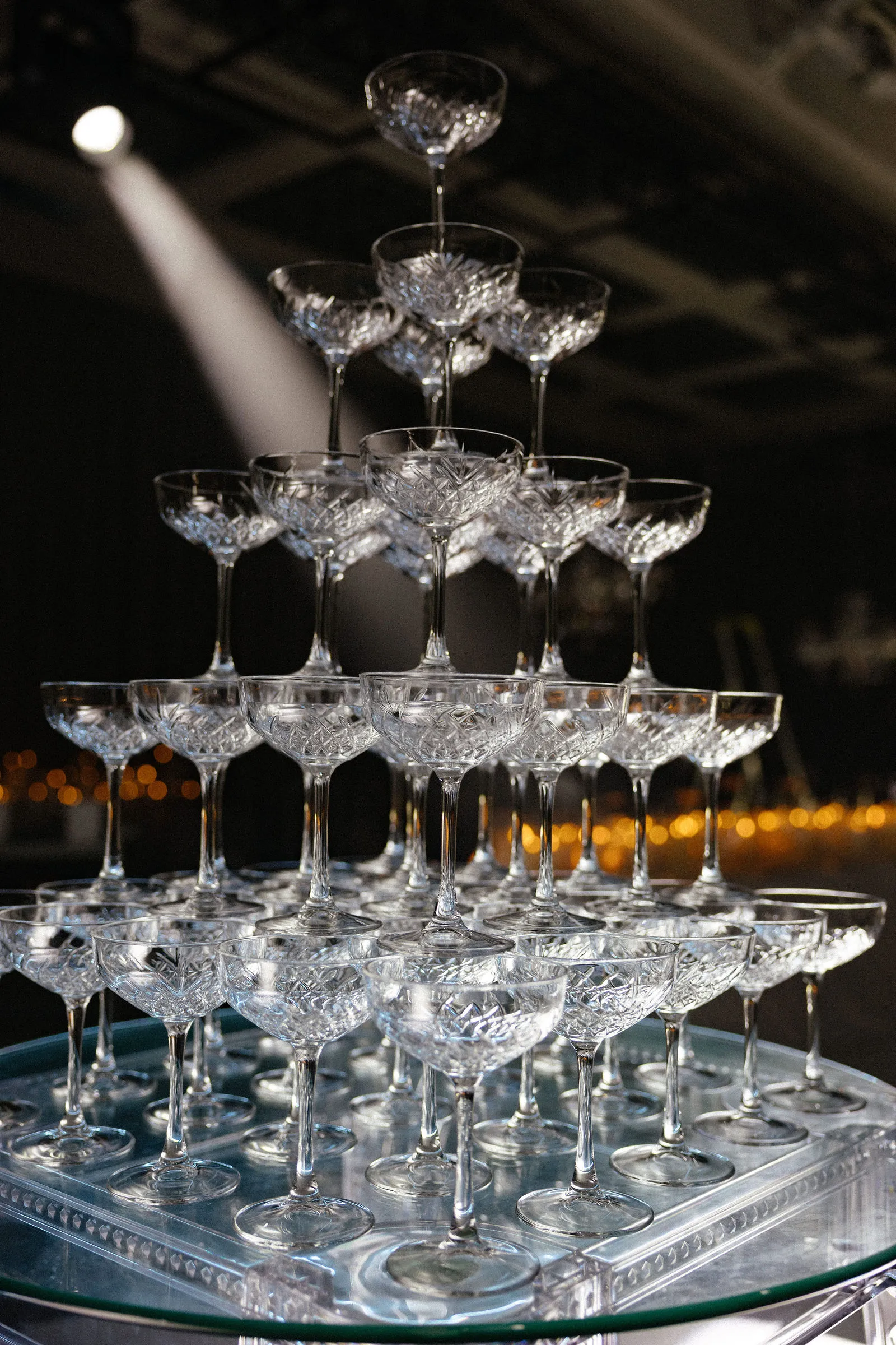 A pyramid stack of empty crystal champagne coupe glasses on a glass table with a dark background and soft lighting.