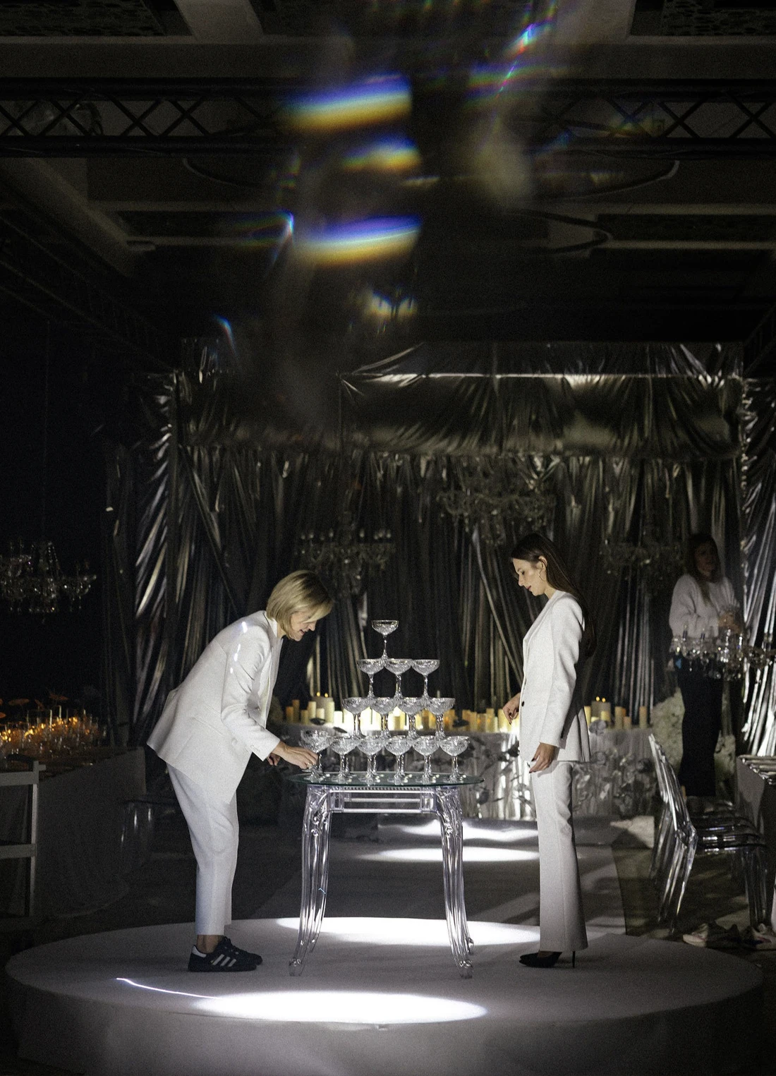 A pyramid stack of empty crystal champagne coupe glasses on a glass table with a dark background and soft lighting.