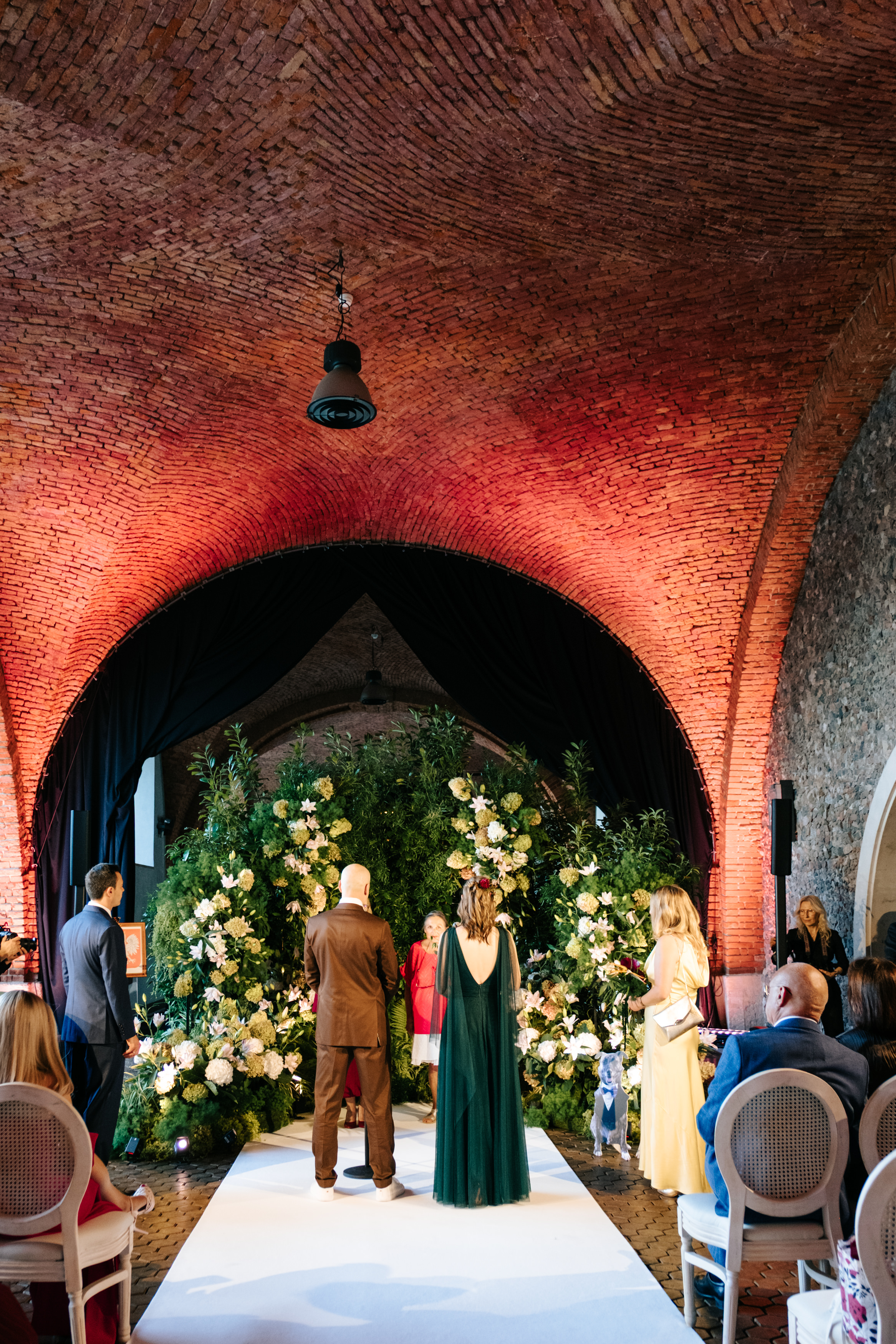 Woman in a backless lace wedding dress standing by a bright window with sheer curtains.