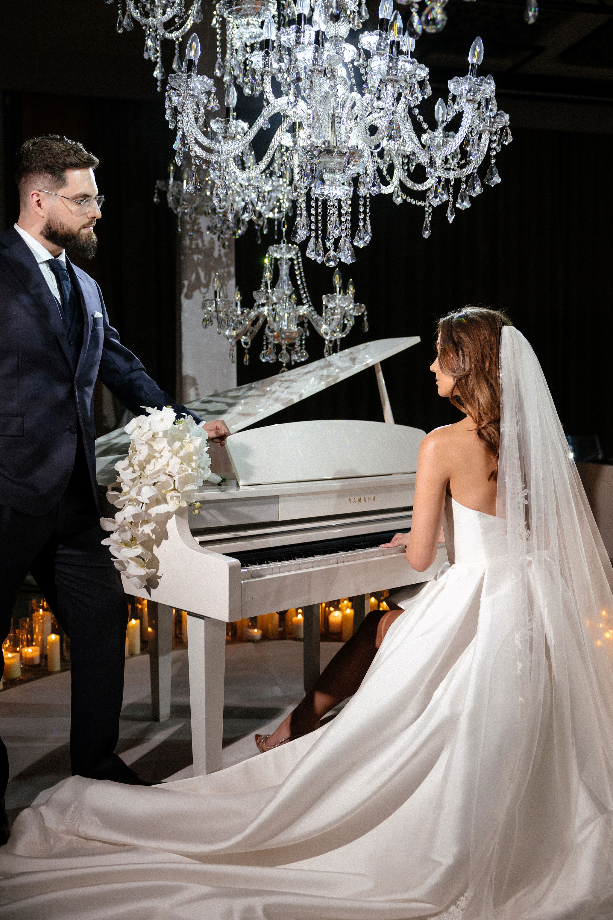Woman in white suit standing beside a pyramid of champagne glasses on an illuminated round platform in a dimly lit, elegant venue with golden curtains and candles in the background.