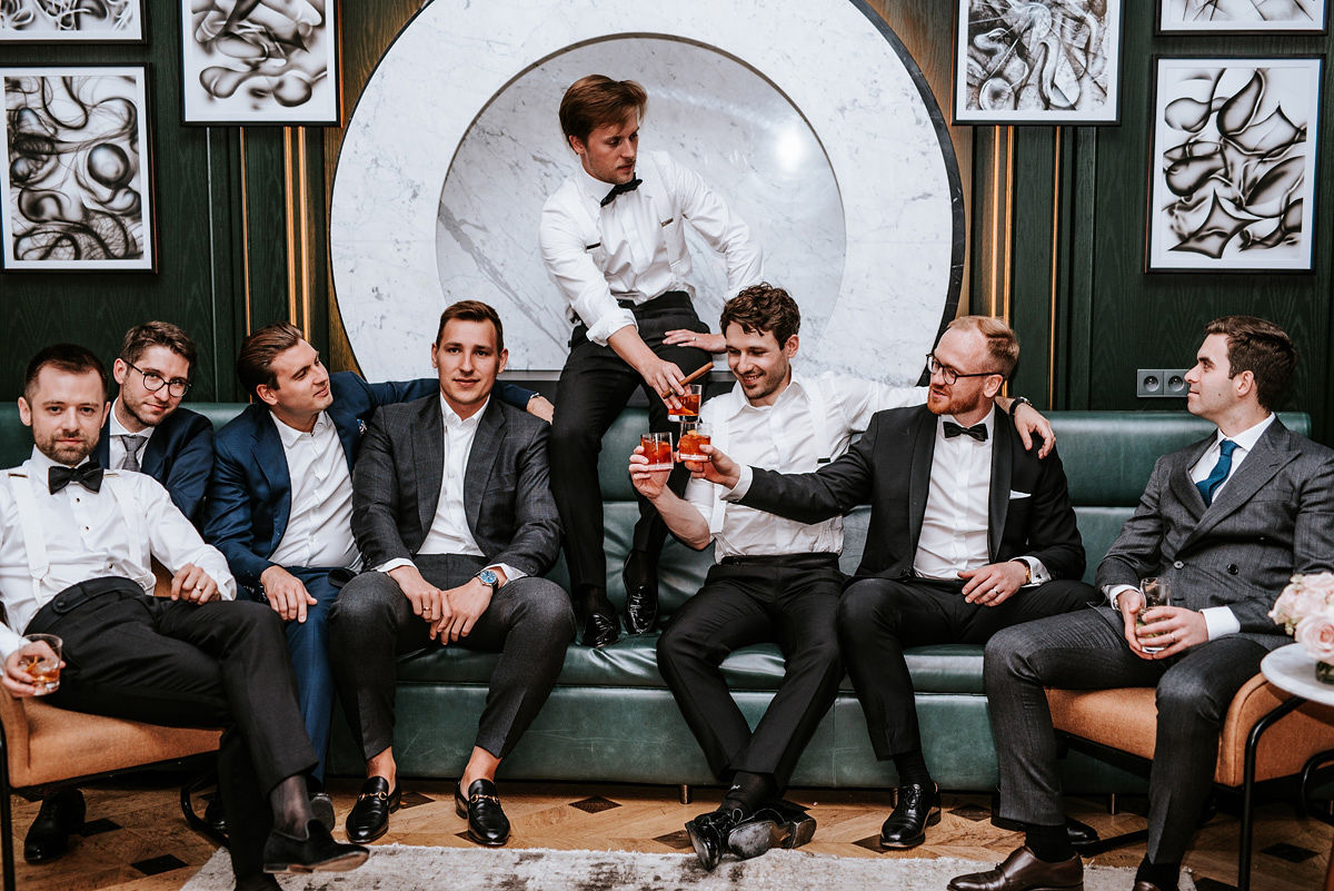 Young man in formal white shirt, black bow tie, and suspenders sitting in a blue armchair holding a phone.