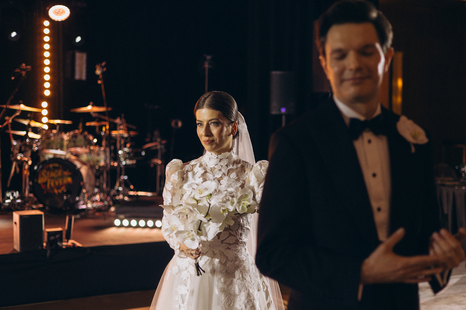 Woman in an elegant lace dress sitting on a round ottoman, putting on strappy heels in a softly lit room.